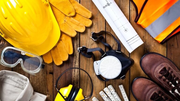 Équipements de sécurité pour le BTP : casque, gants, gilet fluorescent, chaussures et plans de chantier disposés sur une table en bois.